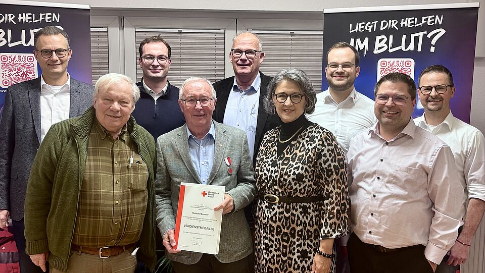 Auf dem Bild sind 9 Personen zu sehen, die frontal augerichtet und lächeln in die Kamera schauen. Eine Person (Herr Reinhard Sommer) hält seine Ehrung in die Kamera "Verdienstmedaille".