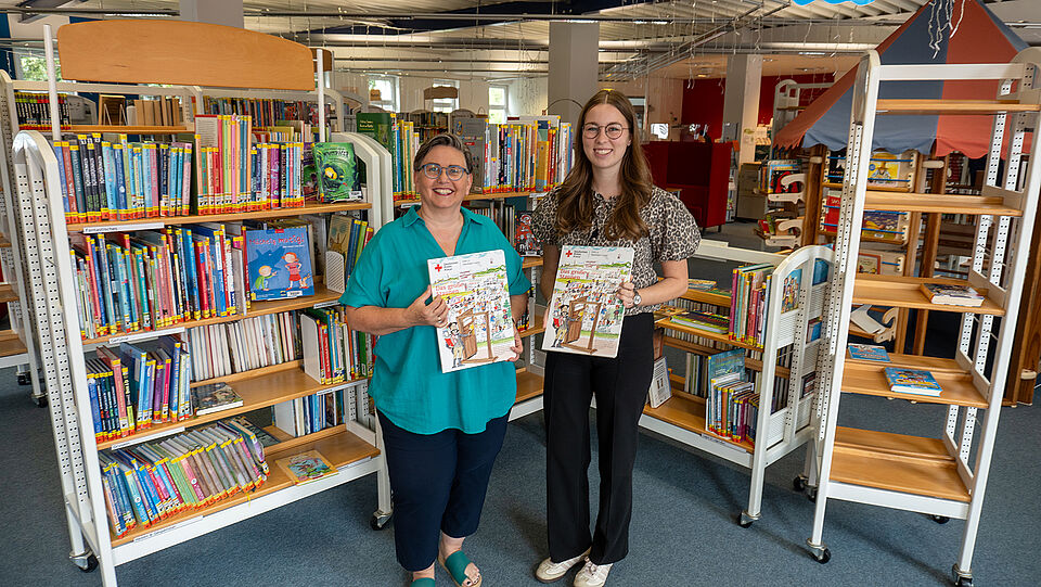 Die beiden Frauen, links die Bibliotheksleiterin Petra Böhler-Winterberg und rechts Werkstudentin Kira Heines, stehen vor Bücherregalen in der Kinder- und Jugendbücherei in Olsberg. Beide haben jeweils ein Wimmelbuch in der Hand und schauen lächelnd in die Kamera. Petra Böhler-Winterberg ist im mittleren Alter, Kira Heines Anfang 20.