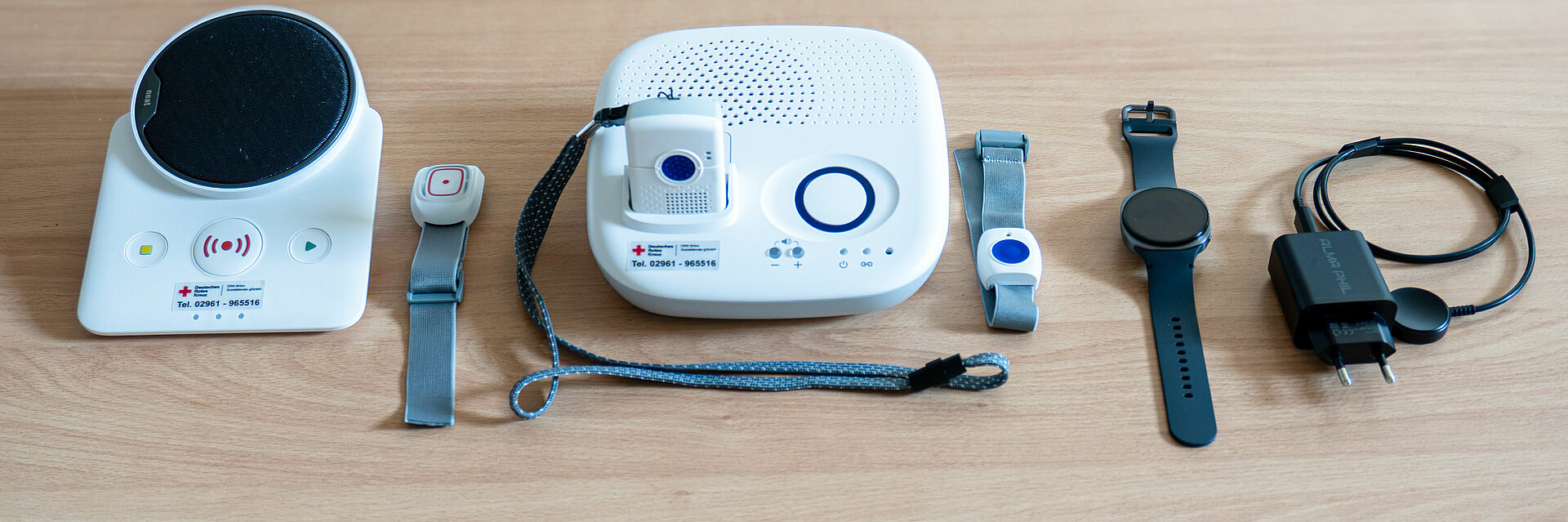 DRK Haustnotrufsysteme Die Basisversion, der Notruf mit mobilem Sender und die Notrufuhr liegen nebeneinander auf einem Tisch