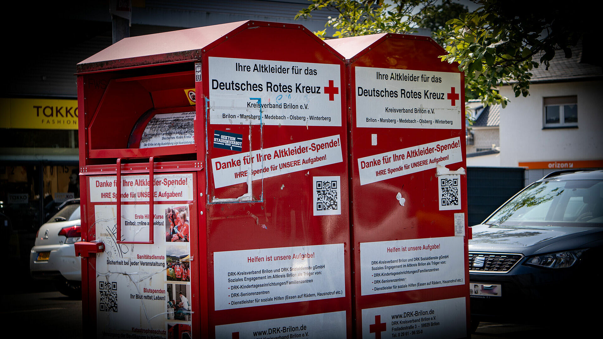 Das Bild zeigt zwei rote Altkleidercontainer des Deutschen Roten Kreuzes (Kreisverband Brilon e.V.), aufgestellt im Freien. Auf den Containern befinden sich große weiße Schilder mit dem roten Kreuz-Logo und Texten in deutscher Sprache, die um Kleiderspenden bitten („Ihre Altkleider für das Deutsches Rotes Kreuz“). Zusätzlich gibt es Informationen zu Verwendungszwecken der Spenden, QR-Codes, Bilder von Hilfsaktionen und eine Liste von Einsatzorten wie Brilon, Marsberg, Medebach, Olsberg und Winterberg. Im Hintergrund sind geparkte Autos, ein Modegeschäft („Takko Fashion“) und ein Gebäude mit orangenem Schild zu sehen.