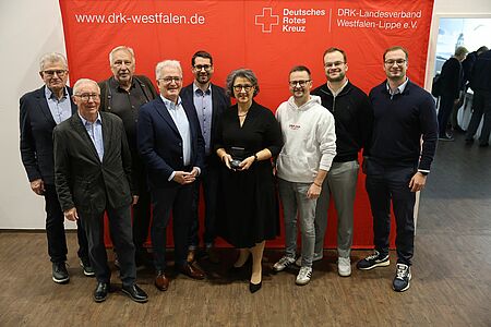 Die sieben Herren und Dame stehen vor einer roten Pressewand des DRK Landesverbands Westfalen-Lippe. Alle Personen sind offiziell gekleidet. Es sind vier ältere Herren und vier jüngere auf dem Bild, die Dame ist ebenfalls etwas älter. Sie steht in der Mitte und hält die Medaille, die übergeben wurde, in der Hand. Alle Personen lächeln und schauen direkt in die Kamera.