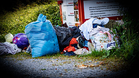 An einer Wiese steht ein Altkleidercontainer des DRK, vor dem einige Säcke mit Kleidung und lose Textilien liegen.